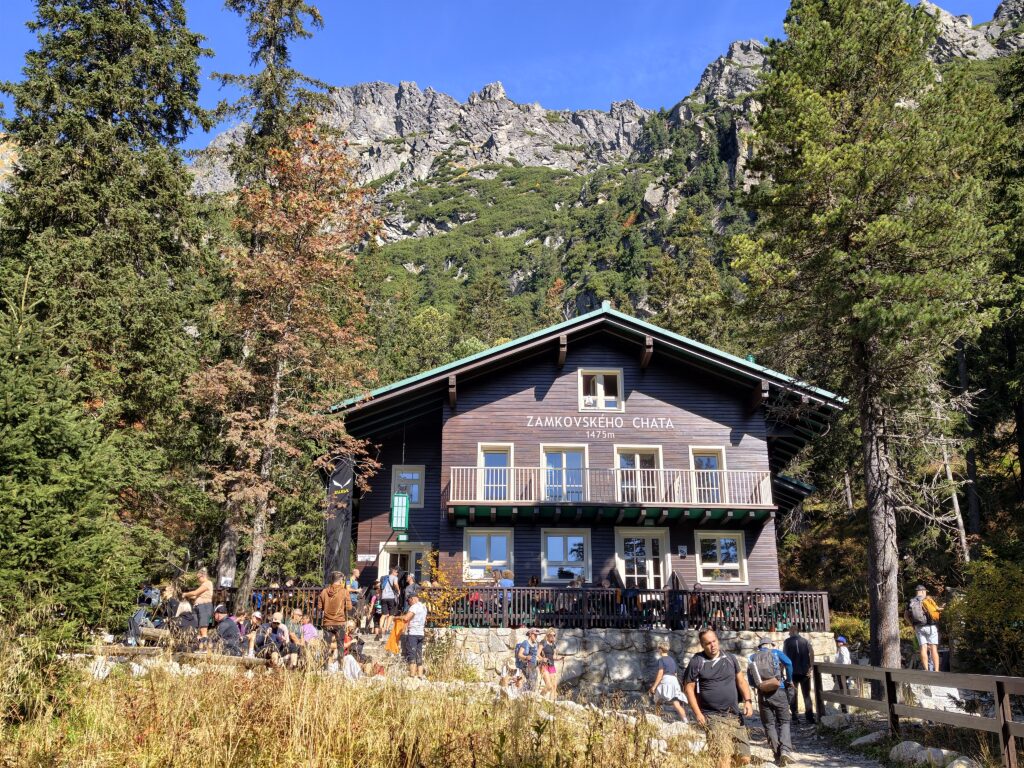 Drewniane schronisko Zamkovského chata (1475 m) z tarasem pełnym turystów i skałami nad dachem, Tatry Wysokie